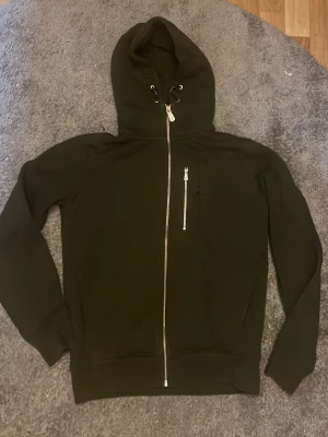 Mörkgrön hoodie med dragkedja - Säljer en mörkgrön hoodie med hel dragkedja framtill och en extra ficka med dragkedja på bröstet. Hoodien har långa ärmar, ribbade muddar och justerbar huva med snören. Perfekt för dig som gillar enkel och stilren design.