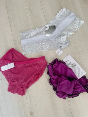 3-pack spets- och satin-trosor - Säljer ett 3-pack trosor i olika stilar: ett par vita spets-trosor med bred kant, ett par ceriserosa spets-trosor och ett par lila satin-trosor med svarta volangkanter. Alla har lappar kvar och är i trendiga modeller. Perfekt för dig som gillar variation och snygga detaljer.