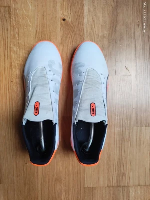 Vita och orangea Puma KING fotbollsskor - Säljer ett par stilrena fotbollsskor från Puma KING-serien. Skorna är vita med orangea detaljer och har en klassisk låg profil. Sidan pryds av den ikoniska Puma-loggan i silver. Ovandelen är i syntetmaterial och skon har en tajt passform för bra bollkontroll.