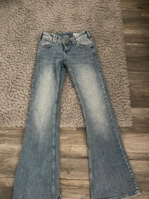 Blå bootcut jeans med spetsdetaljer - Snygga blå bootcut jeans med ljus tvätt och slitningar. Bakfickorna har dekorativa lock med spetsdetaljer och knappstängning. Jeansen har klassisk femficksmodell och är tillverkade i denim med en cool vintagekänsla. KÖPTE NYLIGEN ANVÄNT TVÅ GÅNGER