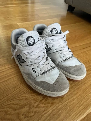 New Balance 550 vita/grå sneakers - Snygga New Balance 550 sneakers i vitt och ljusgrått med mockadetaljer och klassisk logga på plösen. Skorna har lågt skaft, vita skosnören och en stilren retrodesign som passar till det mesta. Perfekta för dig som gillar streetwear och sportig stil.