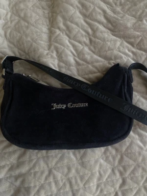Svart axelväska från Juicy Couture - Säljer en svart axelväska från Juicy Couture i mjuk velour med silverfärgad logga framtill. Väskan har en justerbar axelrem med Juicy Couture-tryck och dragkedja upptill. Perfekt för dig som gillar en trendig och klassisk look.