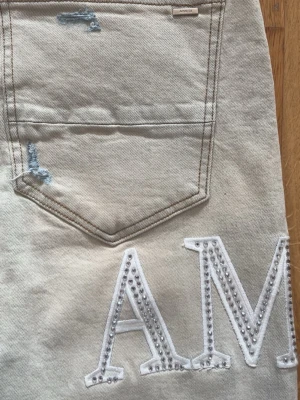 Amiri jeans med vita detaljer - Snygga Amiri jeans med raka ben och slitningar vid fickorna. På baksidan finns stora vita bokstäver med nitar som dekoration. Jeansen har klassiska bakfickor och kontrastsömmar. Perfekta för dig som vill sticka ut med coola detaljer.