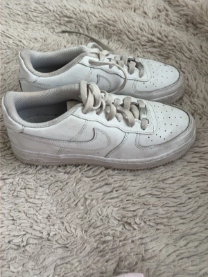 Vita Nike Air Force 1 sneakers - Klassiska vita Nike Air Force 1 sneakers med lågt skaft och perforerad tåbox. Skorna har vit ovandel i läder, vit sula och matchande vita skosnören. Swoosh-logga på sidan och diskret Air-detalj på sulan. Perfekta för en clean och stilren look.