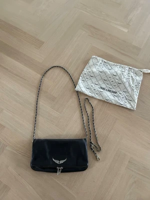 Svart axelväska Zadig & Voltaire - Snygg svart axelväska i läder från Zadig & Voltaire med silverfärgad kedjerem och ikonisk vingdetalj framtill. Om ni ser på bild 3 så är de lite risigt på alla dragkrokar. Men de går lätt att färga till med silverfärgad. Nypris ca 4000