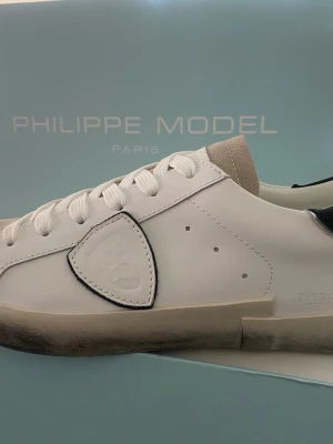 Philippe Model vita sneakers - Snygga vita sneakers från Philippe Model med beige mockadetaljer på tån och svart detalj vid hälen. Skorna har klassisk snörning och en stilren design med märkets logga på sidan. Perfekta för dig som gillar en clean och trendig look.