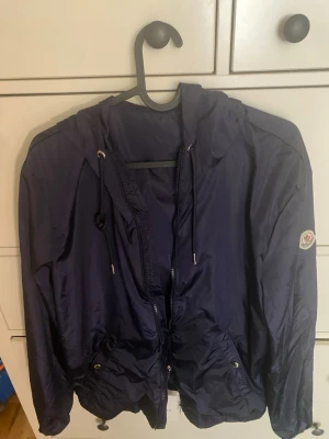 Mörkblå vindjacka med huva - Snygg mörkblå vindjacka med huva och dragsko, hel dragkedja framtill och två fickor med knappstängning. Jackan har ett emblem på ärmen och rödvit detalj på huvan. Tillverkad i ett glansigt, tunt material som passar perfekt för blåsiga dagar.