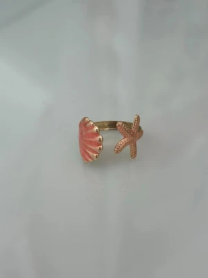 Guldfärgad ring med snäcka och sjöstjärna - Unik guldfärgad ring med öppen design, dekorerad med en rosa snäcka på ena sidan och en matchande sjöstjärna på andra sidan. 