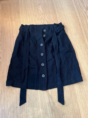 Svart kortkjol med knappar från H&M - Svart kortkjol från H&M L.O.G.G. med knappar framtill och ett matchande bälte i midjan. Kjolen har en enkel och stilren design, perfekt till många olika looks. Tillverkad i ett mjukt material som känns bekvämt.