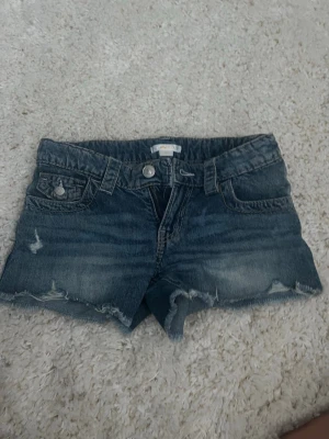 Blå jeansshorts med slitningar - Säljer ett par blå jeansshorts med råa kanter och slitna detaljer. Shortsen har fem fickor, bälteshällor och knappgylf. Perfekta för varma dagar och ger en avslappnad look.pris kan diskuteras 🥰