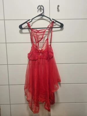 Rött spetsigt babydoll-set - Säljer ett rött babydoll-set i transparent mesh och spets med tunna axelband och dekorativ rosett framtill. Setet har en luftig, asymmetrisk kjoldel och matchande stringtrosa. Perfekt för dig som gillar romantiska detaljer och lätt material.