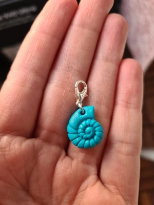 Habdgjord turkos snäcka berlock - Säljer en liten turkosglittrig berlock formad som en snäcka med spiralformad design. Berlocken har ett silverfärgat klolås som gör den enkel att fästa på armband, halsband eller väska. Berlocken är gjord för hand av mig i polymerlera. 