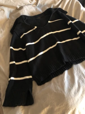 Stickad tröja  - Snygg svart stickad tröja med vita horisontella ränder och v-ringning. Tröjan har långa ärmar och ribbade muddar vid ärmslut och nederkant. Perfekt för dig som gillar en avslappnad och trendig stil.
