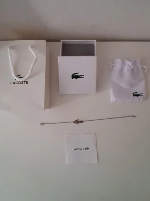 Silverfärgat armband från Lacoste - Snyggt och stilrent armband från Lacoste i silverfärg med den ikoniska krokodilen som detalj. Kedjan är tunn och minimalistisk, perfekt för dig som gillar diskreta accessoarer. Kommer med originalpåse, ask och påse från Lacoste.