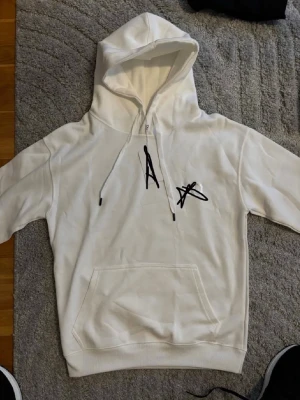 D hoodie vit - Snygg vit hoodie med svart grafiskt tryck på bröstet. Hoodien har huva med dragsko och en stor magficka framtill. Perfekt för en avslappnad och trendig stil.