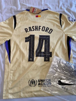 Barcelona Rashford matchtröja guld - Snygg Barcelona matchtröja i guld med svarta detaljer, Rashford och nummer 14 på ryggen. Tröjan har klubbmärke, Spotify-logga och UNHCR-tryck. Korta ärmar, rund hals och tillverkad i lätt, ventilerande material. Perfekt för fotboll eller som supporterplagg.