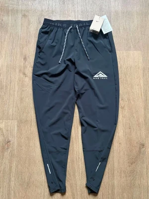 Svarta Nike Trail träningsbyxor  - Svarta träningsbyxor från Nike Trail med dragsko i midjan och fickor på sidorna. Byxorna har smal passform, reflexdetaljer vid bensluten och tryckt logga på låret. Perfekta för en aktiv livsstil och riktigt sköna att ha på sig.