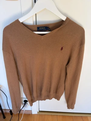 Beige V-ringad tröja Polo Ralph Lauren - Snygg beige V-ringad tröja från Polo Ralph Lauren i mjuk Pima bomull. Tröjan har den klassiska broderade loggan i vinrött på bröstet och ribbade muddar vid ärmslut och nederkant. Perfekt för en clean och stilren look.