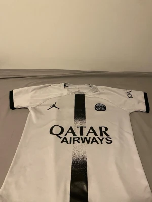 PSG Mbappé matchtröja vit/svart - Säljer en vit PSG matchtröja med svart rand och detaljer, Jordan-logga och Qatar Airways-tryck på bröstet. På ryggen står det Mbappé och nummer 7. Perfekt för dig som älskar fotboll och vill stötta PSG. Materialet är lätt och andas bra.