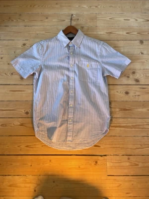 Randig kortärmad skjorta - Snygg kortärmad skjorta med tunna blå och vita ränder. Klassisk krage, knappstängning framtill och bröstficka med liten gul brodyr. Perfekt till jeans eller chinos för en clean look. Använd en gång. 