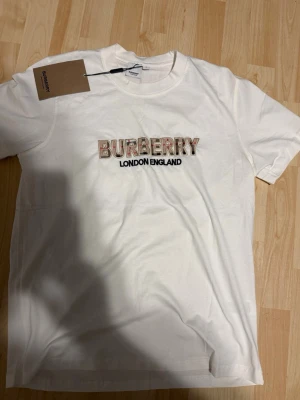 Vit Burberry t-shirt med logga - Säljer en vit t-shirt från Burberry med broderad logga och texten 'London England' på bröstet. T-shirten har klassisk passform, rund hals och korta ärmar. Materialet är mjuk bomull och loggan har ett rutigt mönster i beige, svart och rött.