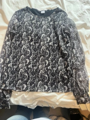 Svart meshblus med ormmönster - Snygg svart meshblus med vitt ormmönster. Blusen har långa ärmar och är gjord i ett tunt, transparent meshmaterial som ger en cool och trendig look. Perfekt att styla med jeans eller kjol för en edgy outfit. Använd många gånger men väldigt bra omhändertagen.