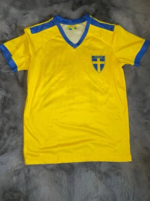 Gul Sverige fotbollströja XS - Gul fotbollströja med blå detaljer från Heja Sverige. Tröjan har V-ringning, blå muddar och axelpartier samt Sveriges landslagsemblem på bröstet. Perfekt för att heja fram Sverige på match eller träning.