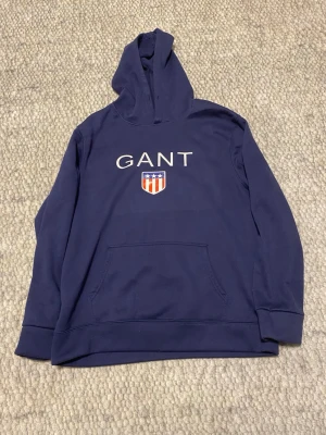 Mörkblå hoodie från GANT - Snygg mörkblå hoodie från GANT med stor logga och sköld på bröstet. Klassisk känguruficka framtill och huva. Tillverkad i mjukt material som passar perfekt till en avslappnad stil. Ribbade muddar vid ärmslut och nederkant.