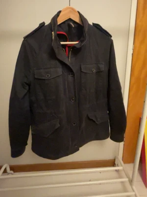 Mörk blå field jacket J Lindberg  - Snygg mörk blå field jacket med fyra stora fickor framtill och axelklaffar. Jackan har hög krage, knapp- och dragkedjestängning samt en rak passform. Perfekt för dig som gillar en stilren och funktionell look. Storlek 40 från J Lindberg. Skriv vid minsta fundering
