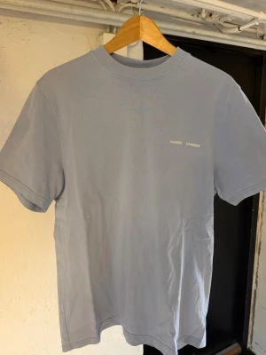 Ljusblå t-shirt från Samsøe Samsøe - En stilren ljusblå t-shirt från Samsøe Samsøe med diskret logga på bröstet. Klassisk passform med rund hals och korta ärmar. Tillverkad i mjuk bomull som känns skön mot huden. Perfekt basplagg till garderoben. Tröjan har blivit lite missfärgad litet precis där bak där du kan se på bilden. 