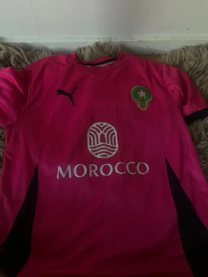 Marocko fotbollströja rosa Puma - Säljer en rosa fotbollströja från Puma med Marockos landslagsemblem på bröstet och trycket 'MOROCCO' framtill. Tröjan har svarta detaljer och är tillverkad i ett lätt, snabbtorkande material. Perfekt för träning eller match!