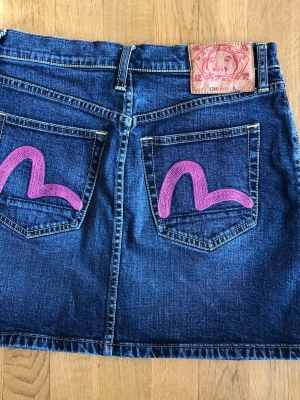 Evisu Jeanskjol med rosa detaljer - Denna jeanskjol från Evisu har en klassisk design med fem fickor och en knappgylf. Kjolen är i en mörkblå denimtvätt med kontrasterande gula sömmar. Baksidan pryds av de ikoniska måsvingelogotyperna i rosa på bakfickorna, samt en stor läderlapp med Evisu-logotypen i midjan.  Längd: 39 cm Bredd linning/midjan: 37 cm Vidd/nederkant: 48 cm