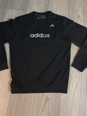 Svart träningssweatshirt från Adidas - Snygg svart sweatshirt från Adidas med klassisk logga på bröstet och rund hals. Tröjan har långa ärmar och ribbade muddar vid ärmslut och nederkant. Perfekt för en sportig och avslappnad stil. OBS: Både Storlek M och möjligtvis L kan passa.