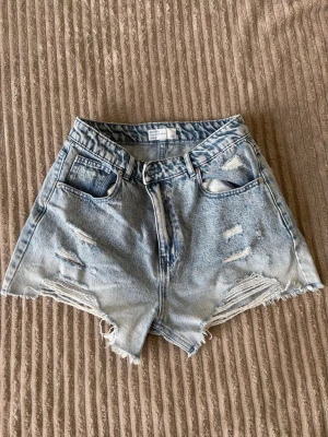 Ljusblå denimshorts med rå kant - Säljer ett par ljusblå denimshorts med hög midja och rå, fransig kant nedtill. Shortsen har två stora bakfickor och klassisk jeanslook. Perfekta för varma dagar och enkel att matcha med olika toppar.