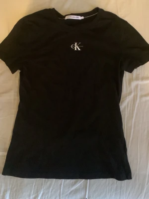 Svart t-shirt från Calvin Klein Jeans - Snygg svart t-shirt från Calvin Klein Jeans med klassisk CK-logga på bröstet. T-shirten har rund hals och korta ärmar, perfekt till alla outfits. Tillverkad i mjuk bomull för skön känsla hela dagen. Själv tycker jag att tröjan i sig känns som en xs. Köpte den då den var lite för liten. 