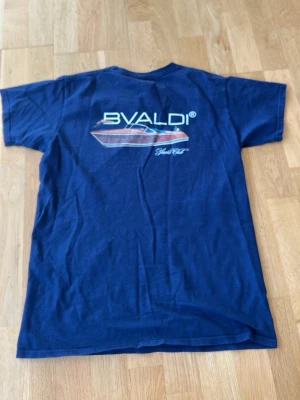 Marinblå BVALDI t-shirt med tryck - Säljer en marinblå t-shirt från BVALDI med coolt båttryck och logga på ryggen samt mindre logga framtill. Klassisk rund hals och korta ärmar. Perfekt för dig som gillar stilrena och marina vibbar.