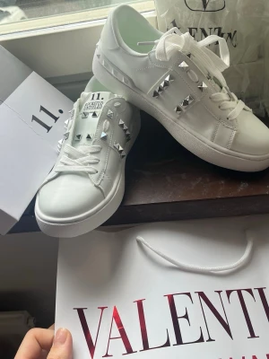 Vita sneakers med nitar från Valentino - Säljer ett par vita sneakers från Valentino med coola silverfärgade nitar på sidorna och ovandelen. Skorna är i läder och har en chunky sula samt vita skosnören. Perfekta för dig som vill sticka ut med en edgy och trendig look.