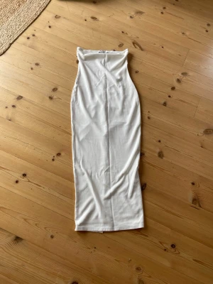 Vit axelbandslös långklänning - Säljer en stilren vit långklänning i mjukt material. Klänningen är axelbandslös och har en kroppsnära passform som framhäver siluetten. Perfekt för sommarens alla tillfällen och enkel att styla med accessoarer.