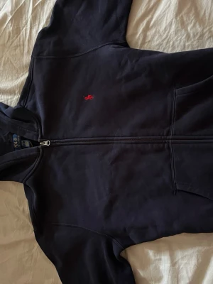 Marinblå hoodie från Polo Ralph Lauren - Snygg marinblå hoodie från Polo Ralph Lauren med dragkedja framtill och klassisk röd logga broderad på bröstet. Hoodien har huva och två fickor framtill. Tillverkad i mjukt bomullsmaterial som är skönt att bära.