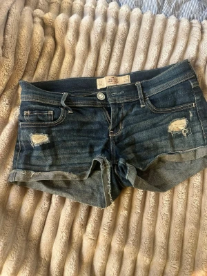 Mörkblå denimshorts från Hollister - Säljer ett par mörkblå denimshorts från Hollister med låg midja och slitna detaljer. Shortsen har klassisk femficksdesign, uppvikta benslut och coola slitningar framtill. 