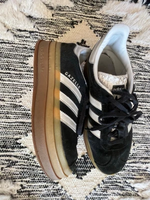 Adidas Gazelle svarta sneakers - Klassiska Adidas Gazelle sneakers i svart mocka med vita detaljer och de ikoniska tre ränderna på sidan. Skorna har en brun gummisula och svart snörning. Insidan är vit och plösen har ett mönstrat tyg. Perfekta för en sportig och stilren look.
