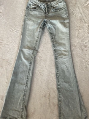 Ljusblå bootcut jeans - Säljer ett par ljusblå bootcut jeans med klassisk femficksdesign och snygga detaljer på bakfickorna. Jeansen har ganska så navellämgd vid midjan och är tillverkade i stretchigt denimtyg för skön passform. Perfekta för dig som gillar retrovibbar och vill ha ett par jeans som sticker ut.
