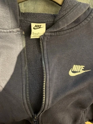Mörkblå Nike zip hoodie med huva - Snygg mörkblå hoodie från Nike med dragkedja framtill och klassisk vit logga på bröstet. Hoodien har huva och mjukt fleecefoder på insidan, samt ribbade muddar vid ärmar och nederkant. Perfekt för en chill och sportig look.