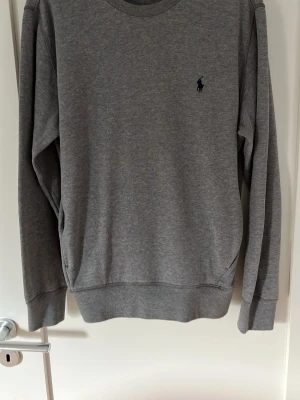 Grå sweatshirt från Ralph Lauren - Klassisk grå sweatshirt från Ralph Lauren med rund halsringning och diskret mörkblå logga broderad på bröstet. Tröjan har långa ärmar och ribbade muddar vid ärmslut och nederkant. Perfekt för en avslappnad och stilren look. Tröjan är heller aldrig använd! Nypris på cirka 2000kr