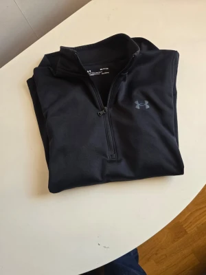 Svart halvzip från Under Armour - Säljer en svart långärmad tröja från Under Armour med halv dragkedja framtill och diskret logga på bröstet. Perfekt för löpning eller gym. Tillverkad i ett mjukt och stretchigt funktionsmaterial som andas.