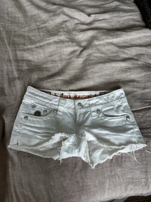 Vita jeansshorts från Rock Revival - Säljer ett par vita jeansshorts från Rock Revival med råa kanter och snygga detaljerade bakfickor. Shortsen har silverfärgade knappar och nitar, samt broderade fickor både fram och bak. Perfekta för sommaren!