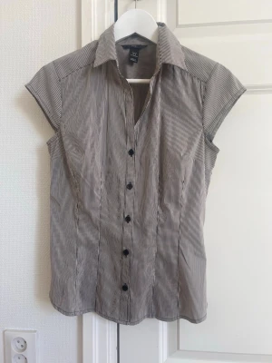 Randig kortärmad blus med krage - Snygg blus med smala gråa och vita ränder, klassisk krage och knäppning framtill. Modellen är figurnära och har korta ärmar. Köpt secondhand från HM