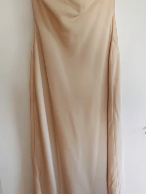 Beige långklänning med snörning - Stilren beige långklänning i mjukt material med snörning vid halsringningen. Klänningen har en rak passform och är ärmlös, vilket ger en enkel och modern look. Perfekt för dig som gillar minimalistisk stil.