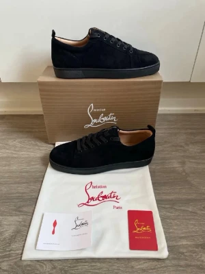 Svarta sneakers från Christian Louboutin - Stilrena svarta sneakers från Christian Louboutin i mocka med svarta skosnören och diskret logga på insidan. Låg modell med klassisk siluett och svart sula. Perfekta för dig som gillar exklusiv och tidlös design.