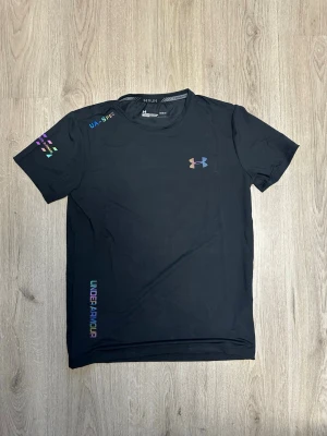 Svart tränings-tshirt Under Armour - Snygg svart tränings-tshirt från Under Armour med färgglada loggor och detaljer. Tillverkad i funktionsmaterial som andas, perfekt för löpning eller gym. Rund halsringning och korta ärmar. UA-logga på bröstet och text på ärmen och sidan.